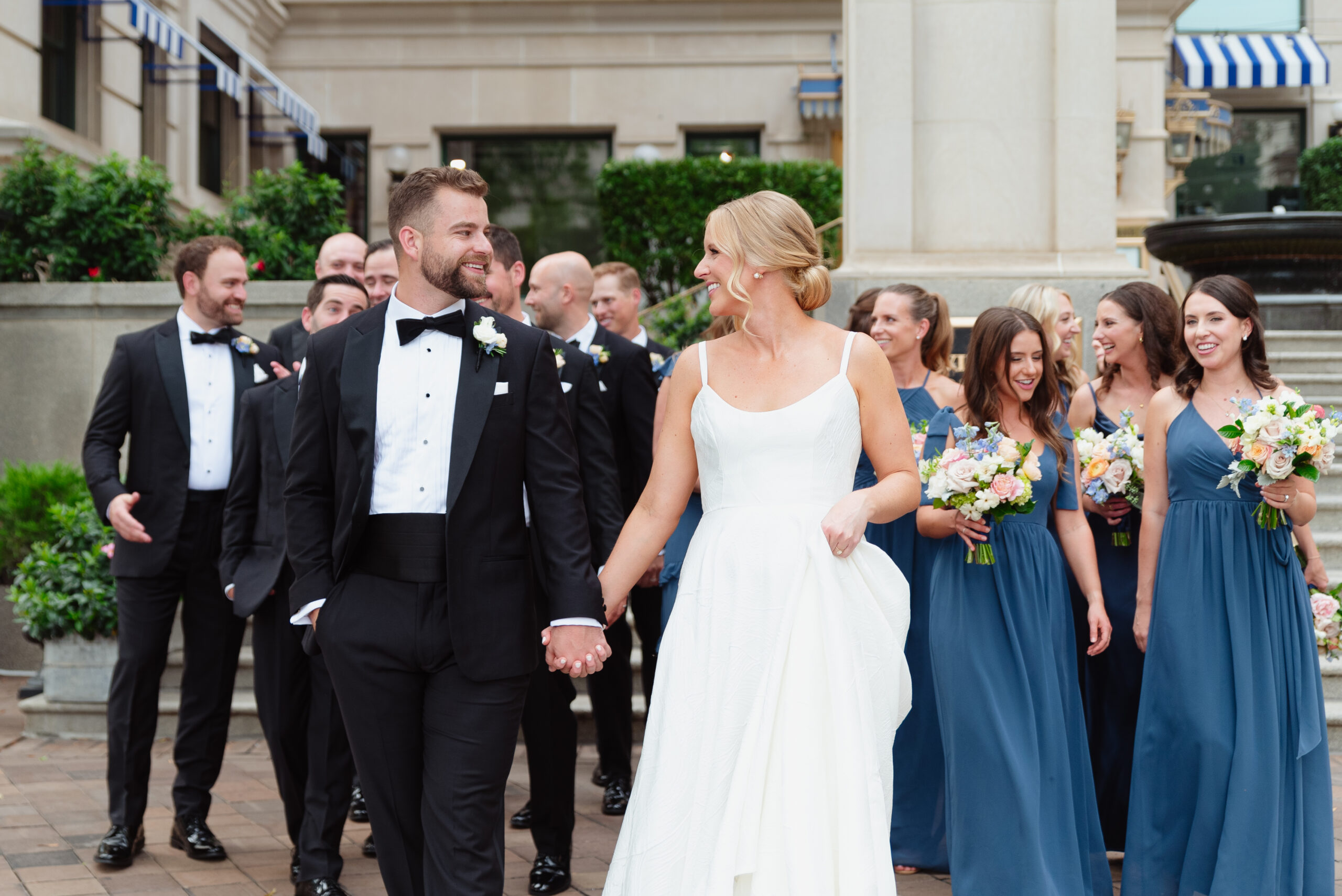 A romantic wedding portrait taken at the Willard InterContinental in Washington, D.C. The couple stands in front of the grand entrance of the hotel, with its elegant columns and classic architectural details. The bride wears a stunning white gown with lace accents, while the groom is dressed in a sharp black tuxedo. Soft, natural light highlights the couple as they share a quiet moment, with the iconic D.C. building providing a sophisticated backdrop.